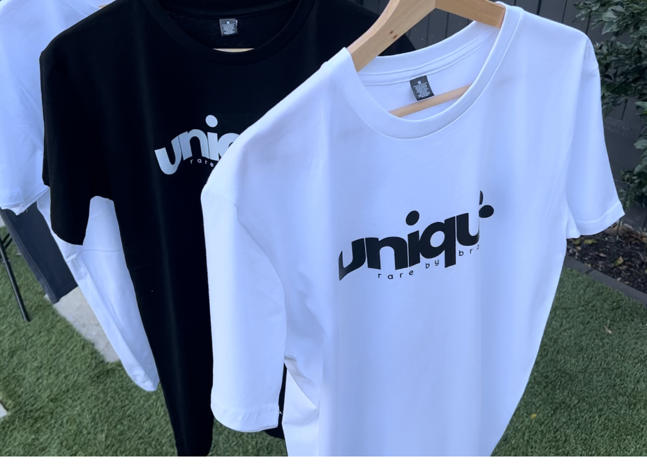 Alternate image of two mens crew neck tshirts, one in black and one in white, hanging on coat hangers outside in a garden..