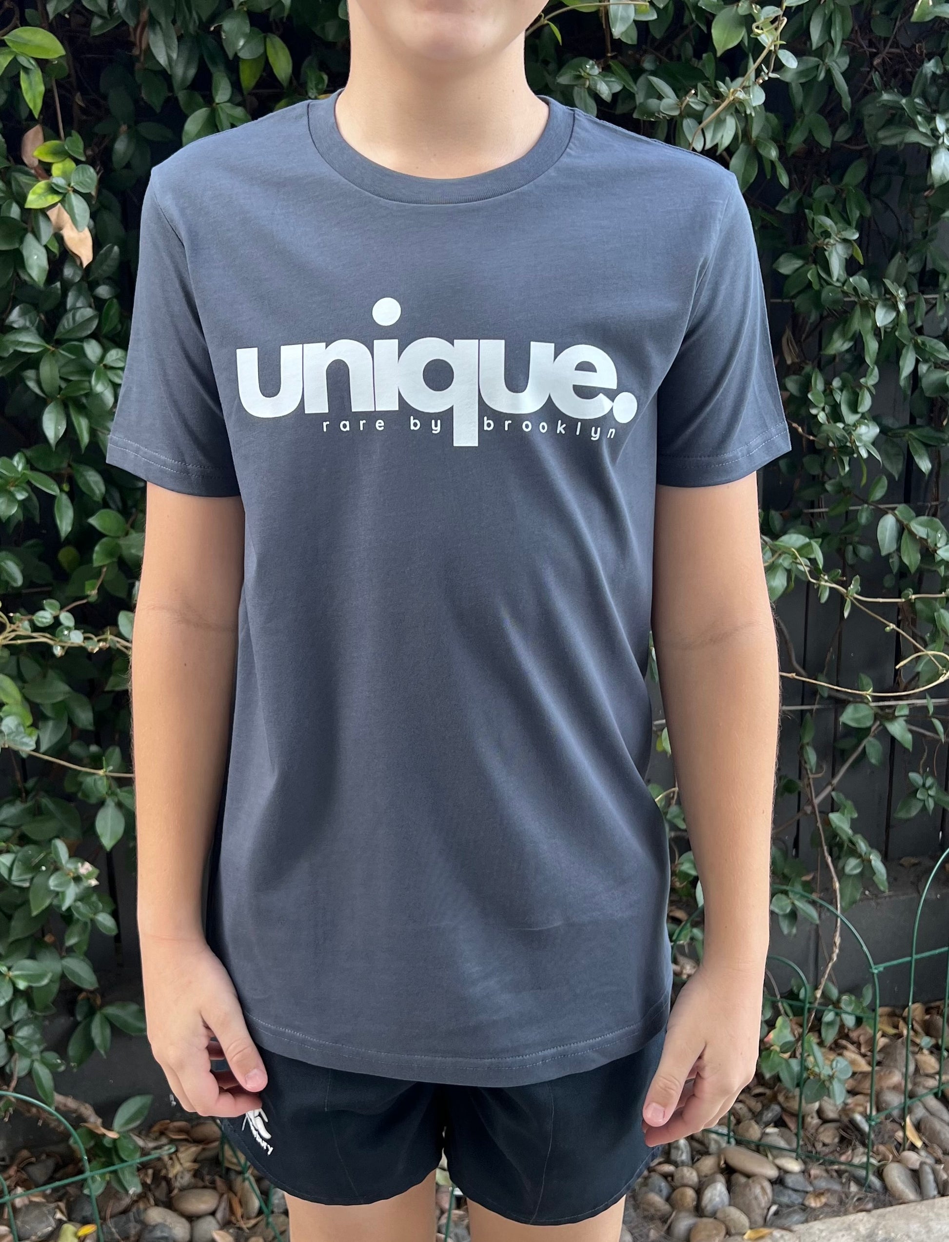 Petrol blue, crew neck, classic fit, youth tshirt. The words 'Unique. Rare by Brooklyn' at featured across the front of the shirt in white print. Image shows youth standing straight with greenery behind.